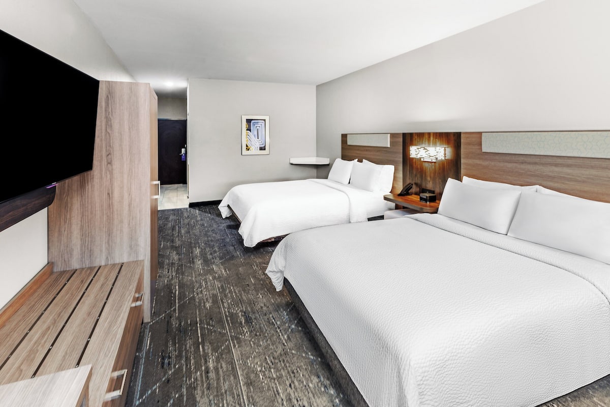 Ein Hotelzimmer mit zwei Betten, einem Flachbildfernseher, einer Wand mit Holzvertäfelung und einem kleinen Nachttisch mit Lampe.