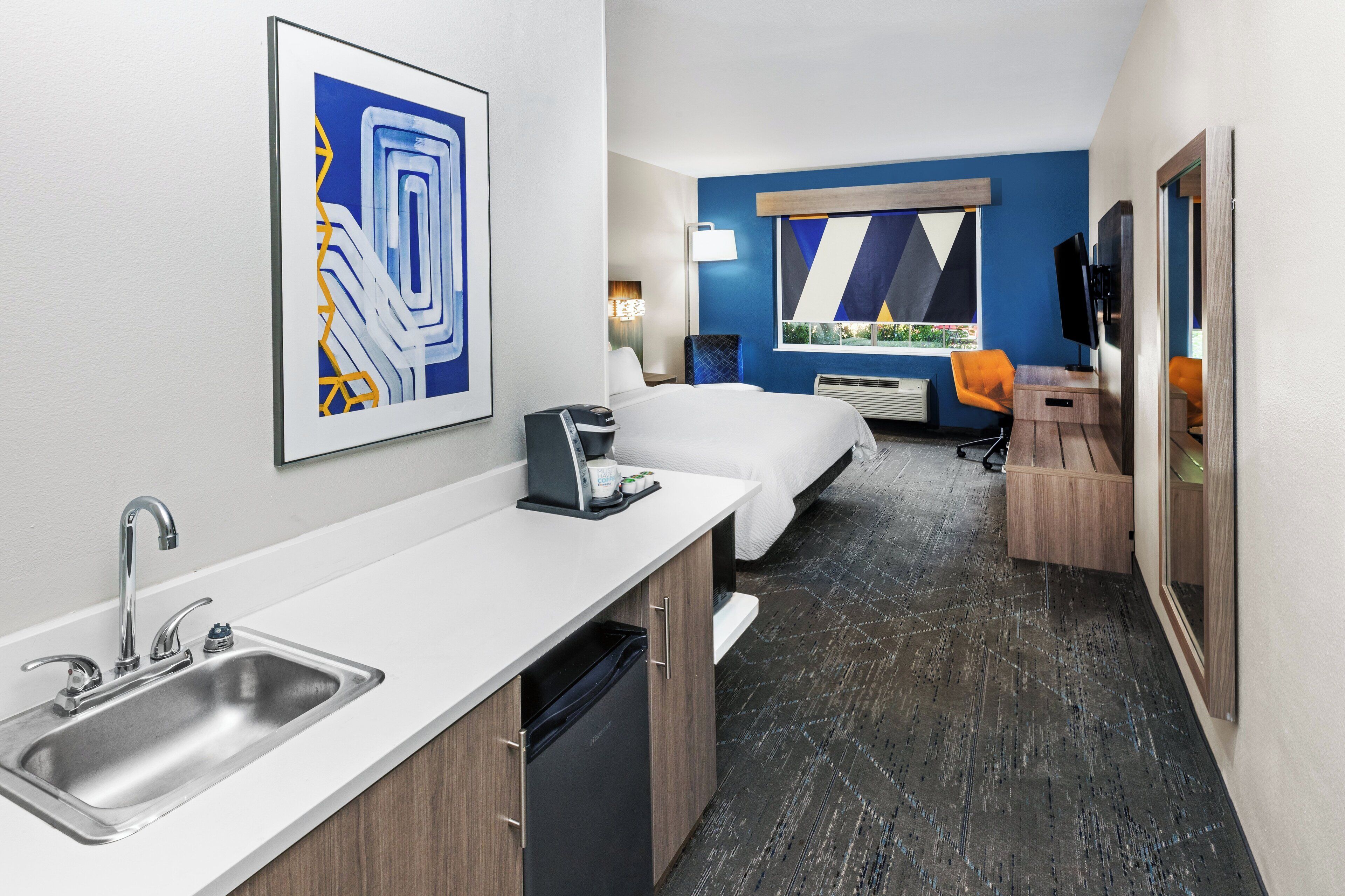 A modern hotel room with a kitchenette, a bed, a desk, and a large window with a view.
