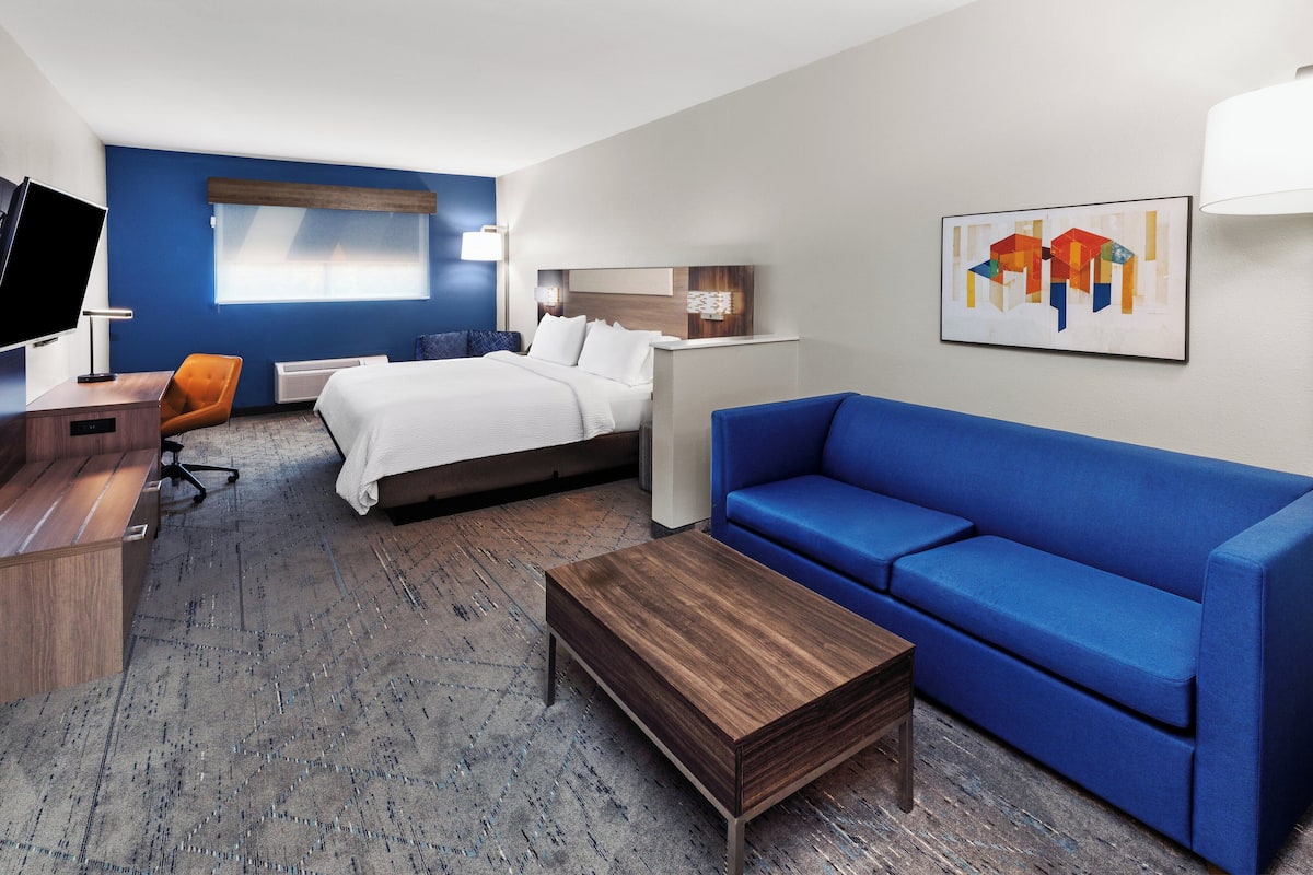 Ein Hotelzimmer mit einem Bett, einem blauen Sofa, einem Holztisch, einem Schreibtisch mit Fernseher und einem Wandbild.