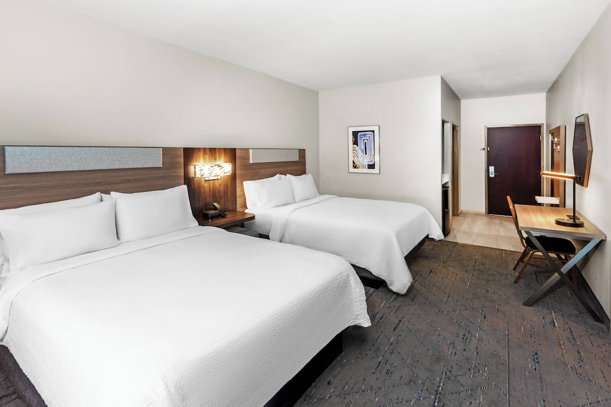 Ein Hotelzimmer mit zwei Betten, einem Schreibtisch, einem Stuhl, einem Nachttisch und einer Wandlampe.