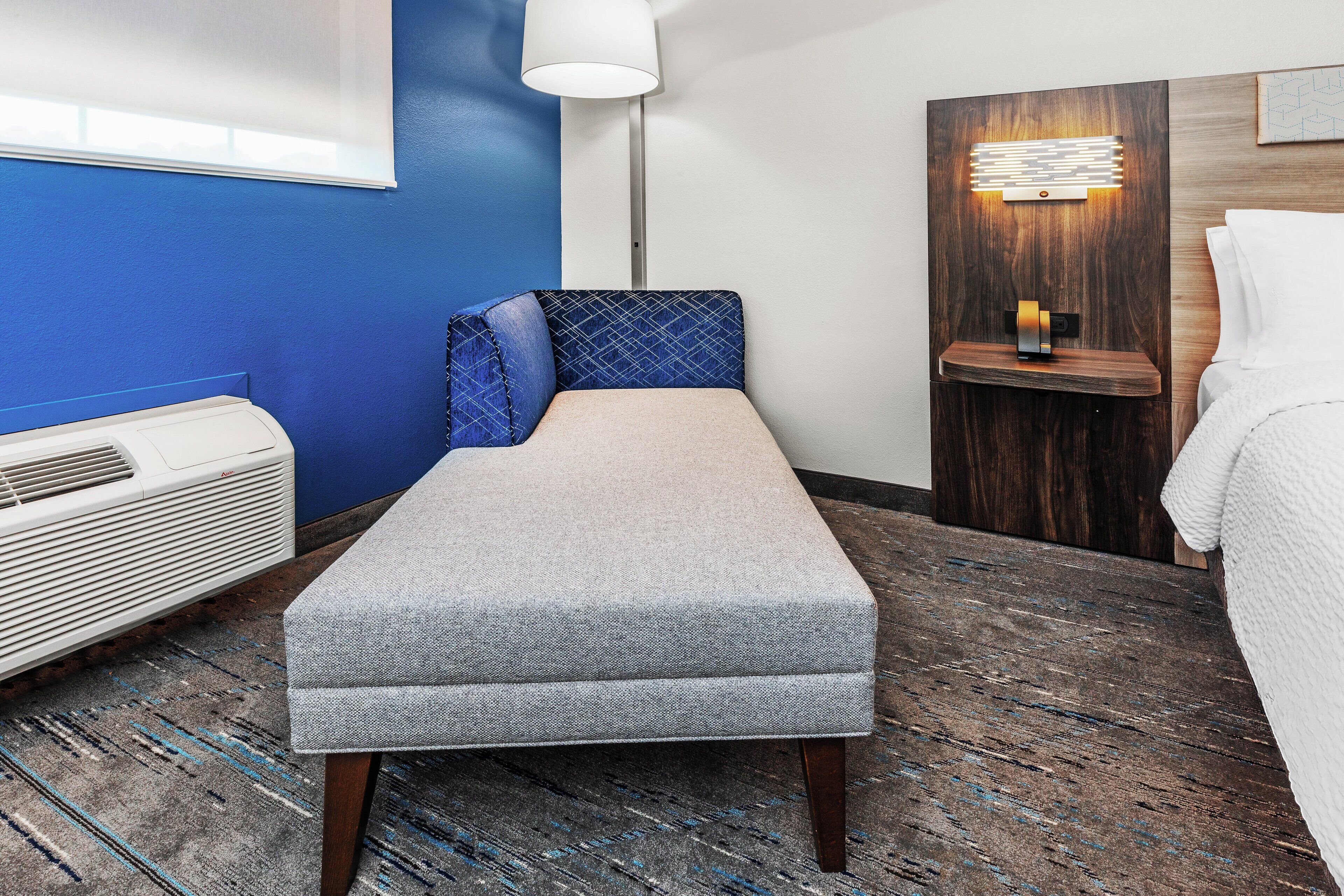 A small, neatly arranged room with a bench, a bedside table with a lamp, and a bed with white bedding.
