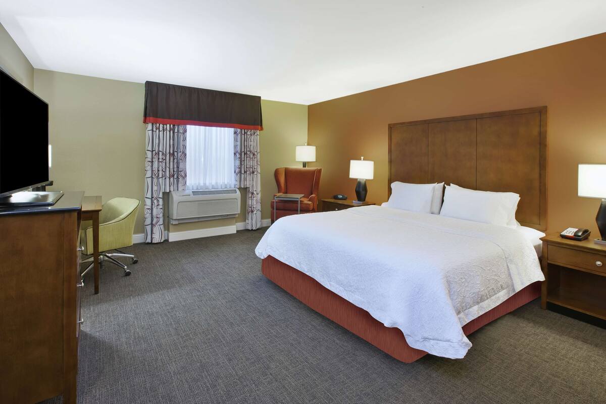 A hotel room with a large bed, a desk with a flat-screen TV, a chair, two bedside lamps, and a window with curtains.