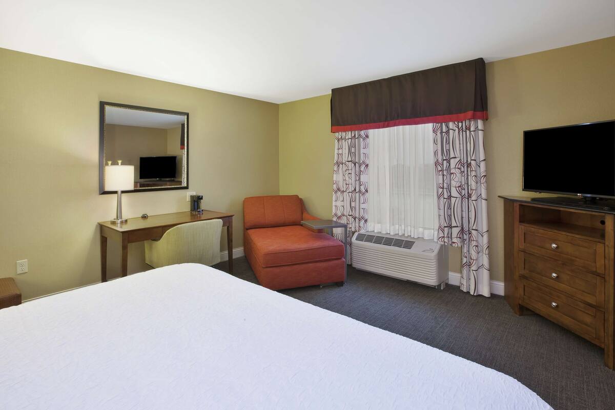 A hotel room with a bed, a desk with a lamp, an orange chair, a television, and a window with curtains.