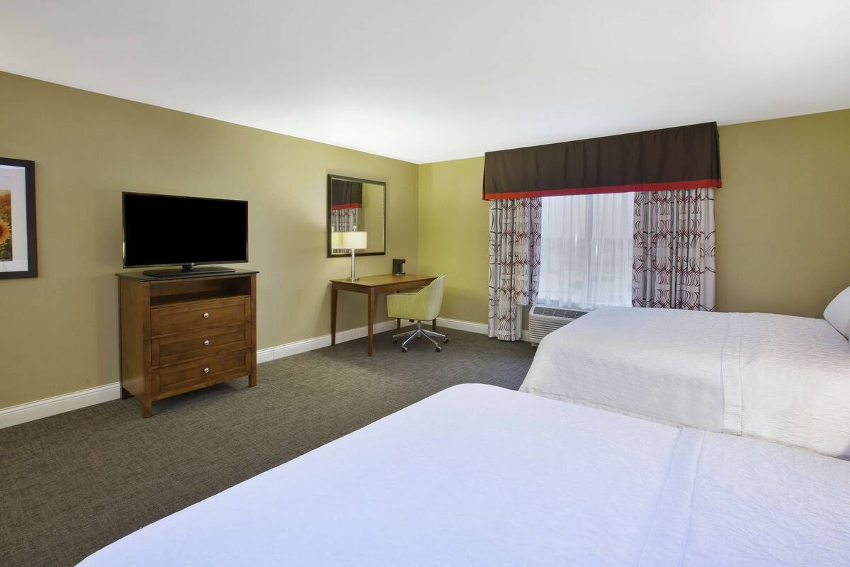 A hotel room with two beds, a desk, a television, and a window with curtains.