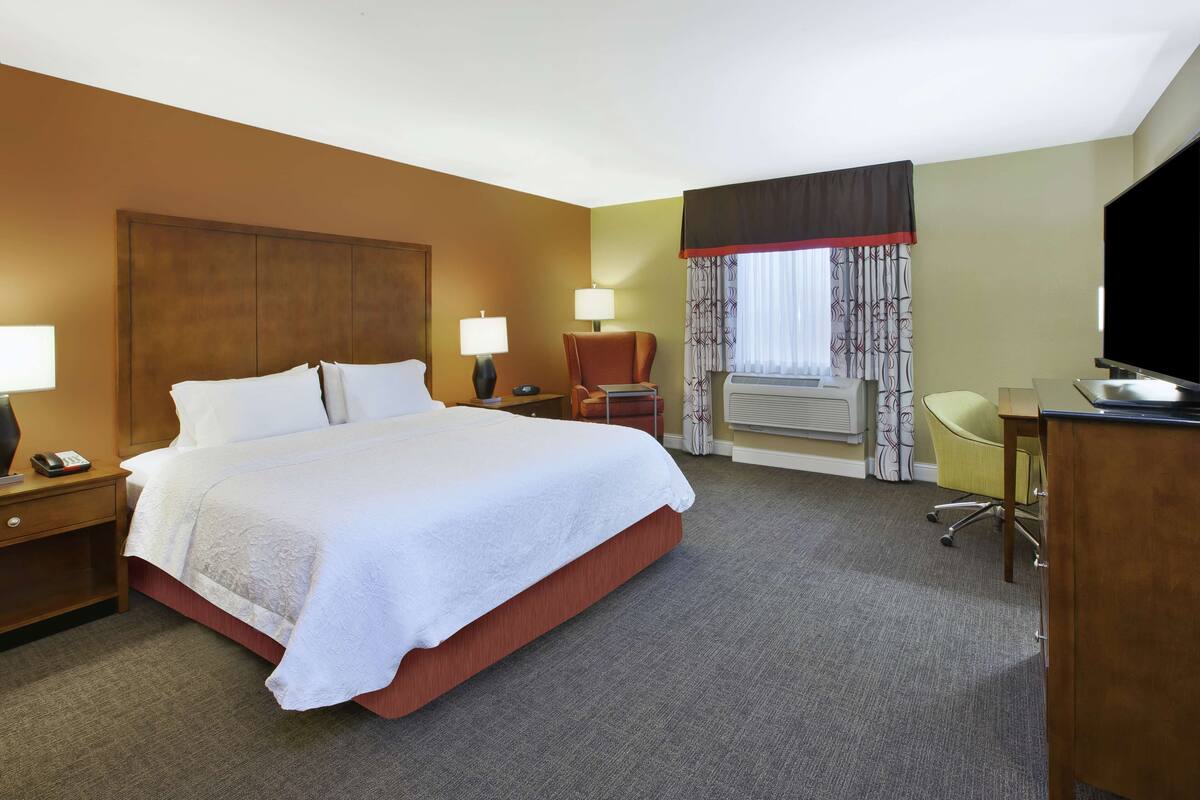 A hotel room with a large bed, bedside tables with lamps, a desk with a chair, a television, and a window with curtains.