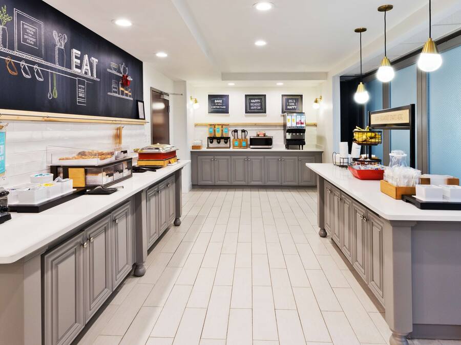 Una cocina moderna con una pared de pizarra que muestra dibujos y texto relacionados con la comida, una isla central con diversos alimentos y una encimera con cafeteras y condimentos.