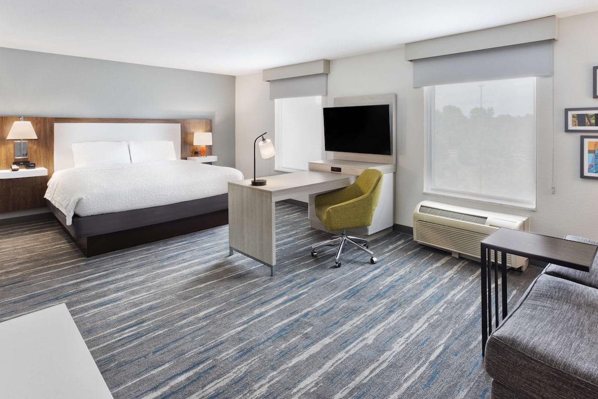 A hotel room with a bed, desk, chair, TV, and air conditioning unit.
