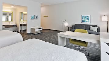Habitación de hotel con dos camas, un escritorio, un sofá y una mesita. Se observa un espejo, una lámpara y obras de arte en las paredes.
