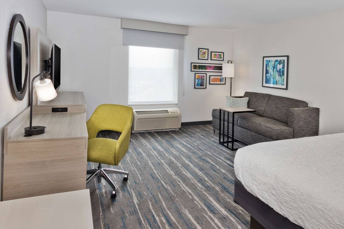 A hotel room with a bed, a desk with a lamp, a chair, a sofa, a TV, and a window with blinds.