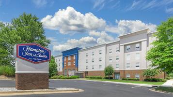 A Hampton Inn & Suites sign with a brick base, a blue and red sign, and a modern building with multiple windows and a blue roof in the background.
