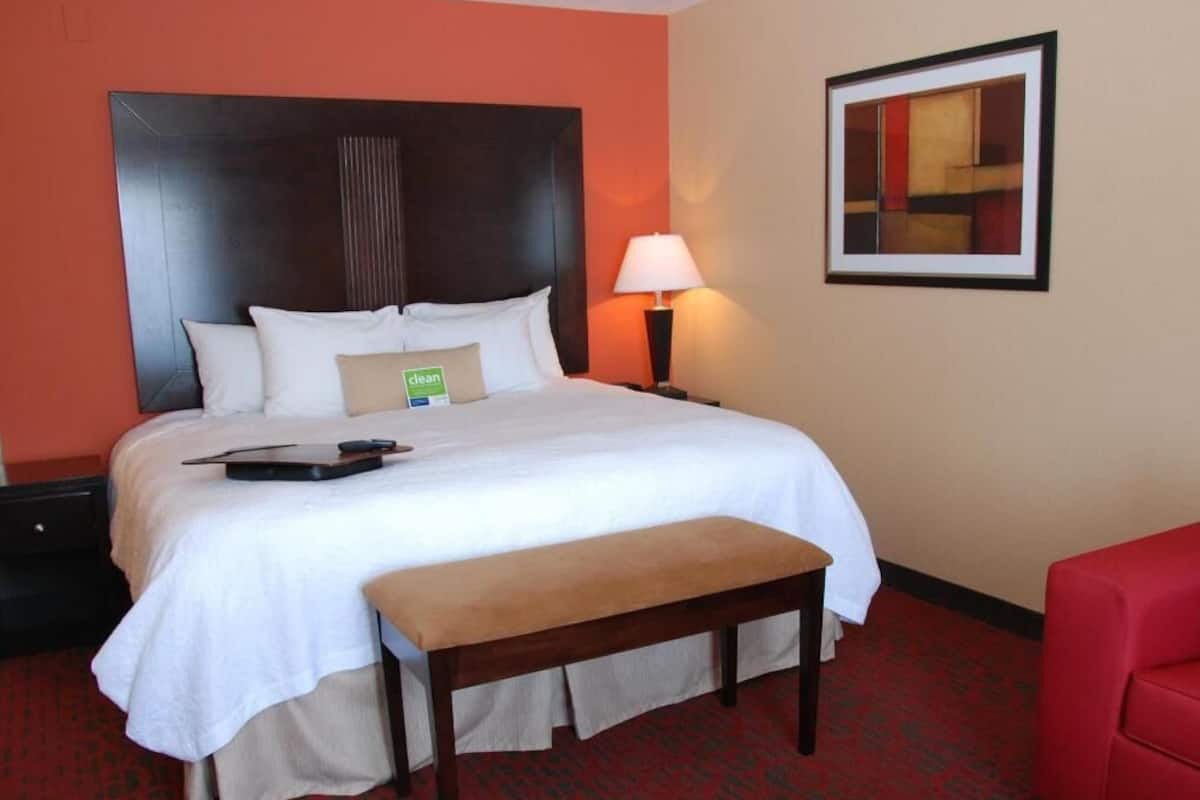 A hotel room with a bed, a bench, a nightstand with a lamp, and a framed picture on the wall.