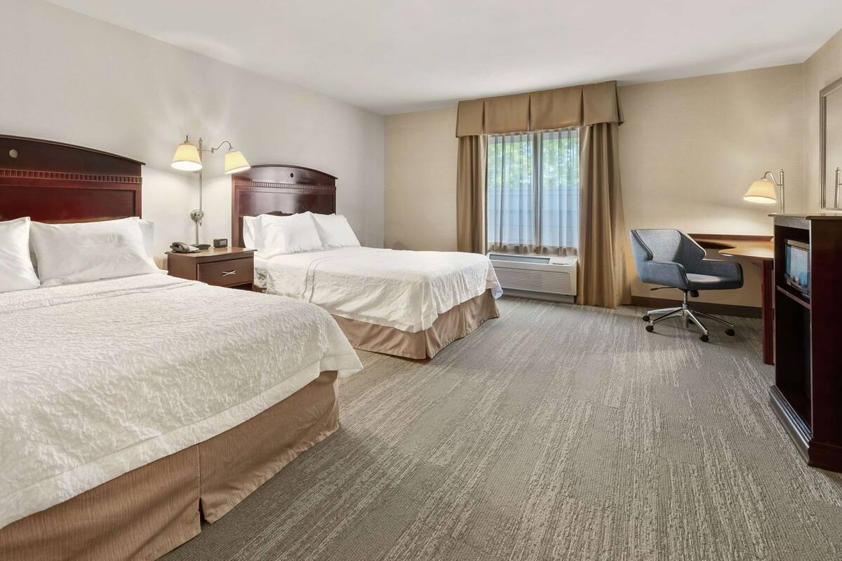 Quarto de hotel com duas camas, uma escrivaninha, uma cadeira, um abajur e uma janela com cortinas.