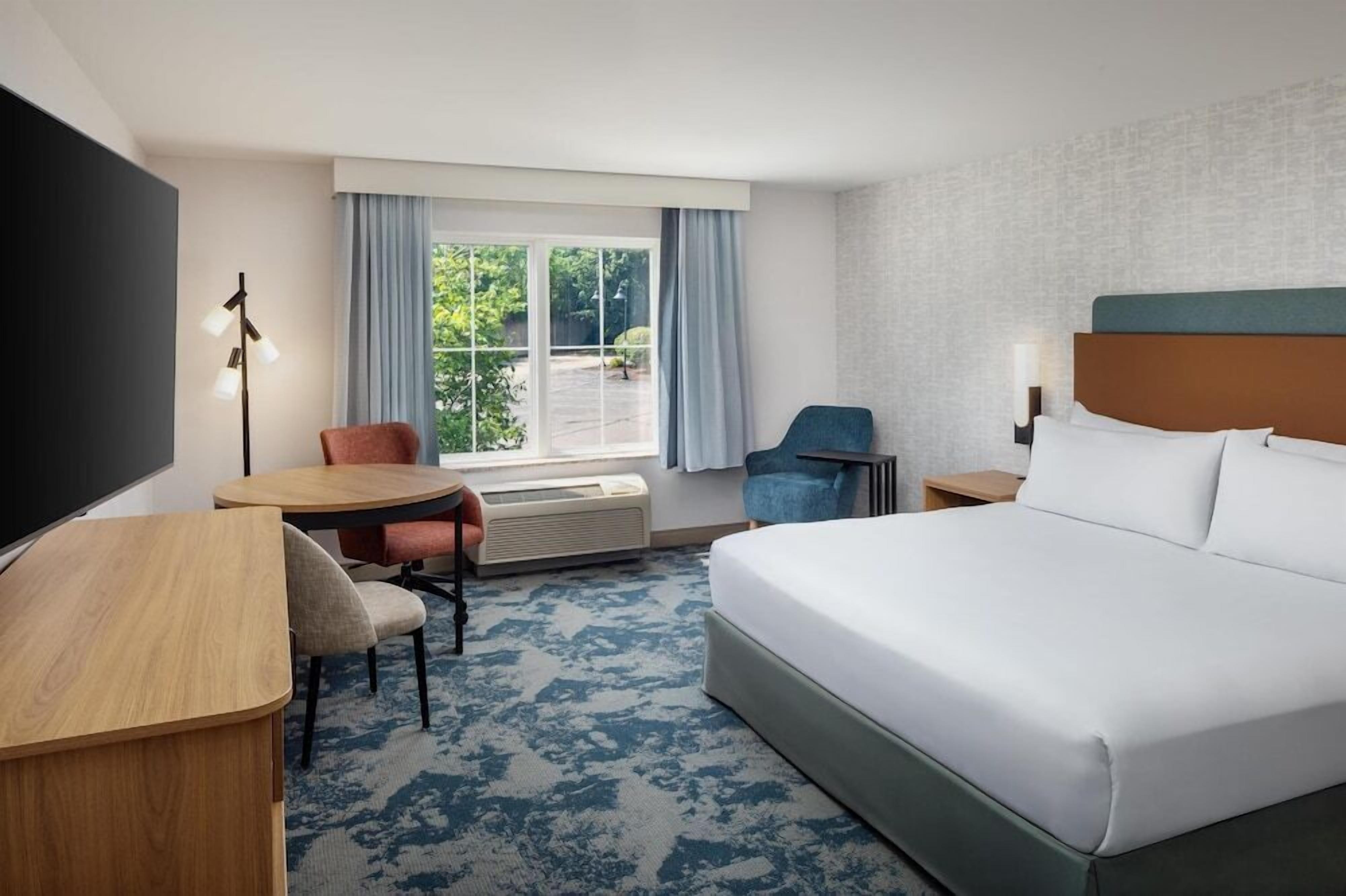 Ein Hotelzimmer mit Bett, Schreibtisch, Stuhl, Fernseher und einem Fenster mit Vorhängen, mit Blick auf Grünflächen.