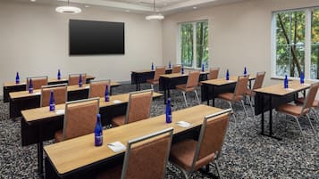 A conference room with multiple tables and chairs, a projector screen, and windows with a view of trees.