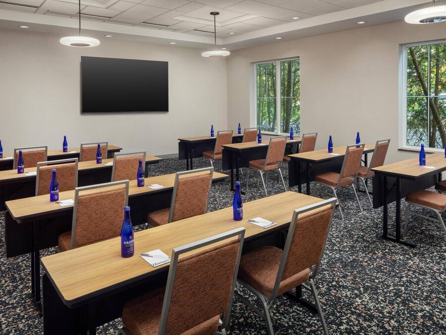 A conference room with multiple tables and chairs, a projector screen, and windows with a view of trees.