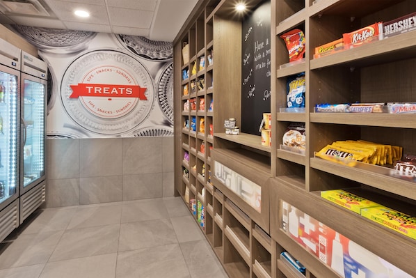 Interior de una tienda con estantes llenos de dulces y refrigerios, un expositor refrigerado y una pared con un diseño circular y la palabra 'TREATES'.