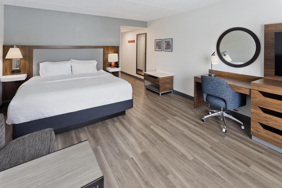 A hotel room with a large bed, a desk with a chair, a mirror, and a TV.