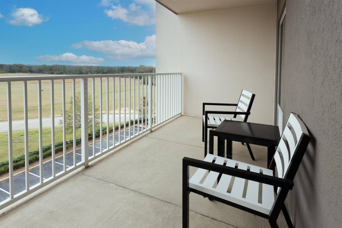 A balcony with a table and chairs, a view of a parking lot and greenery, and a clear sky.