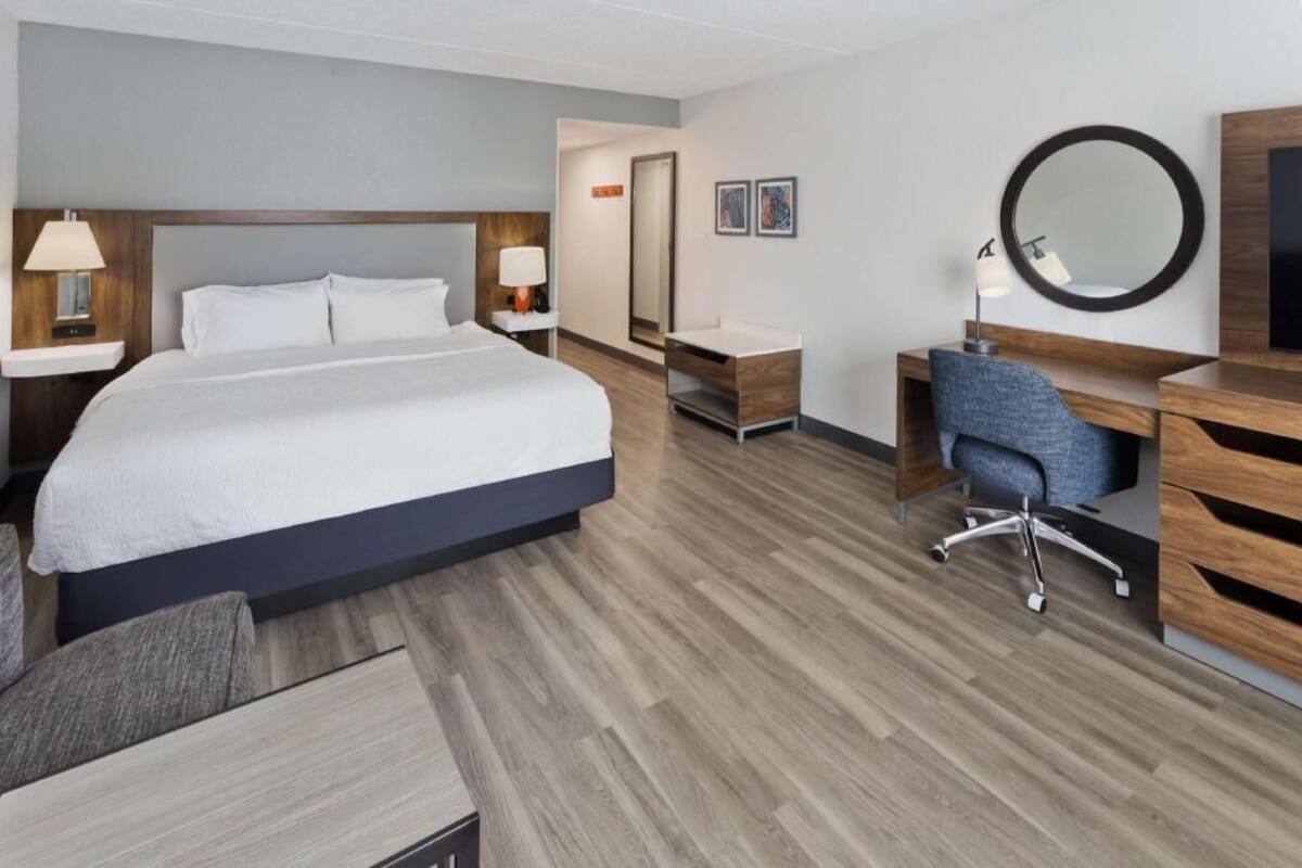 A hotel room with a large bed, a desk, a chair, a mirror, and a TV.