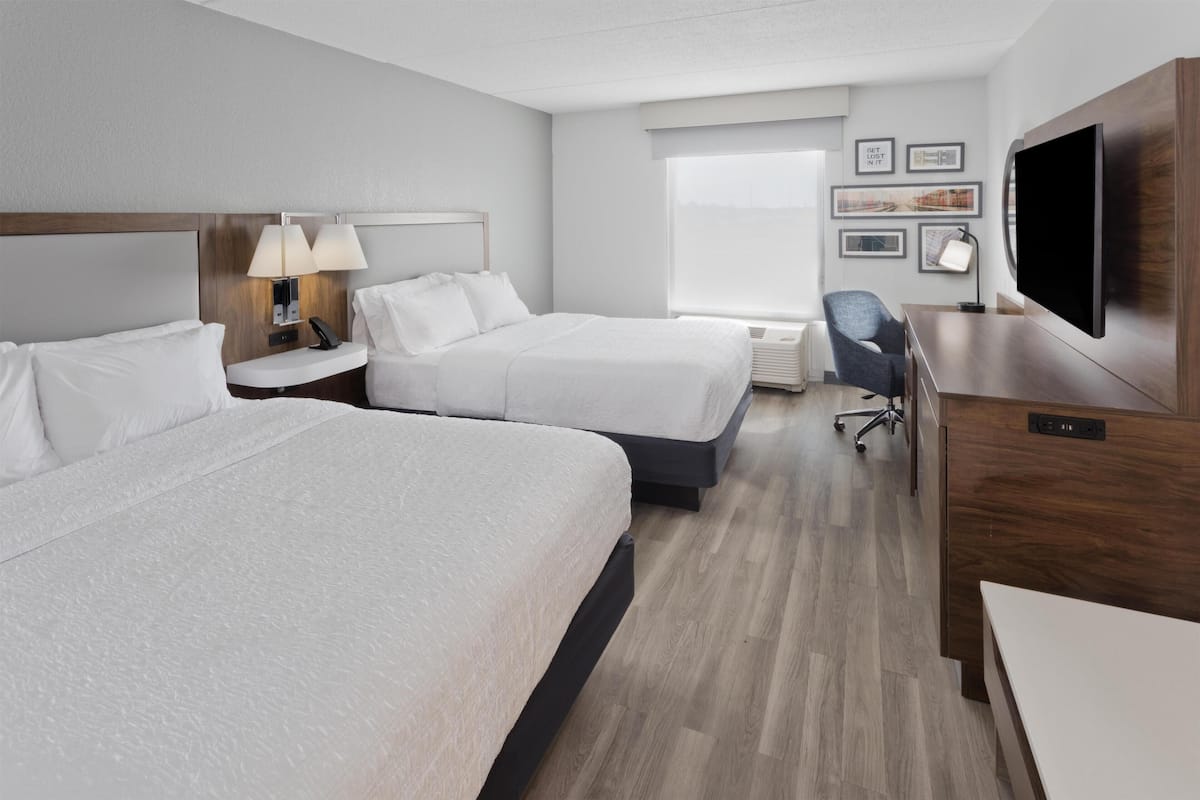 A hotel room with two beds, a desk, a chair, a TV, and a window with a view.