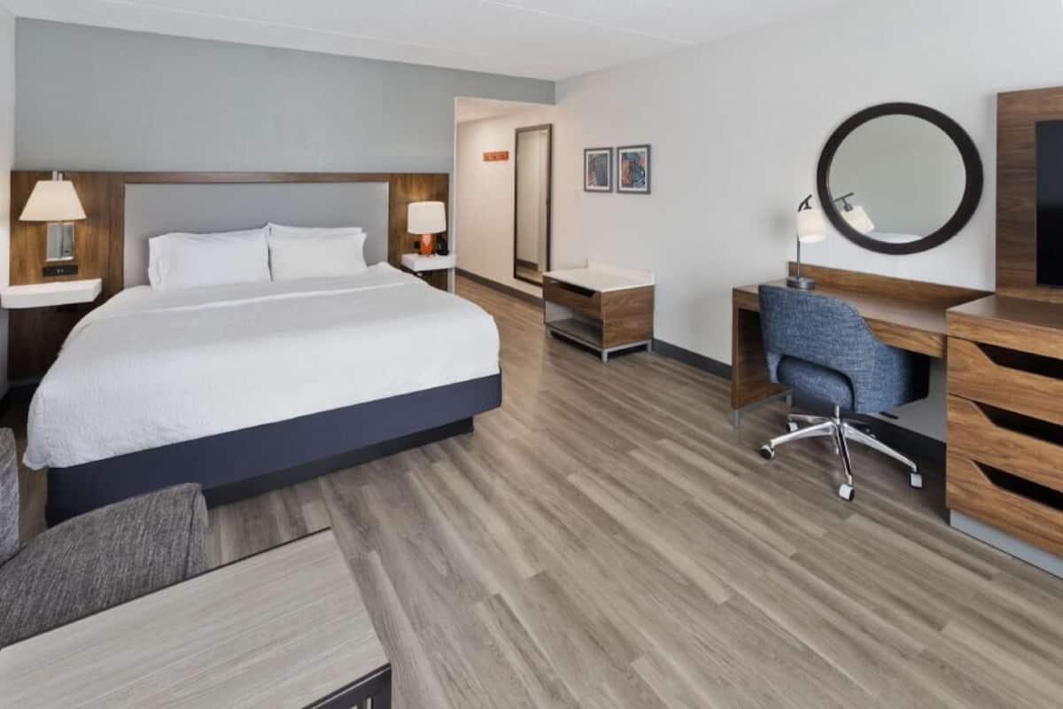 A hotel room with a large bed, a desk, a chair, a mirror, and a TV.