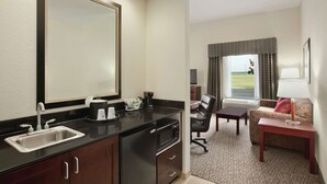 A compact hotel room with a kitchenette, a large mirror, a sofa, and a view of the outdoors.