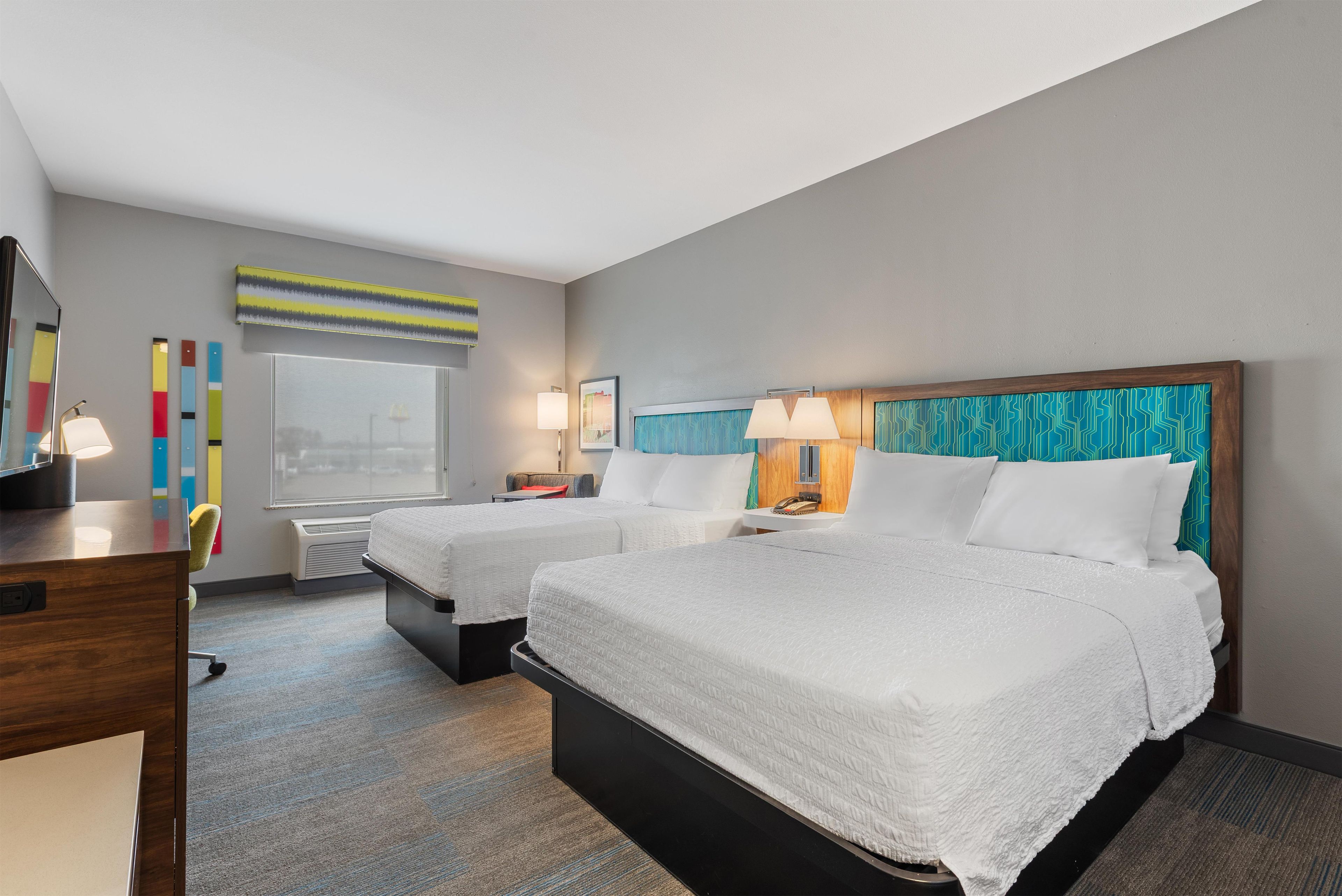 Habitación de hotel con dos camas, un escritorio, una silla, un ventanal y una pared con obras de arte coloridas.