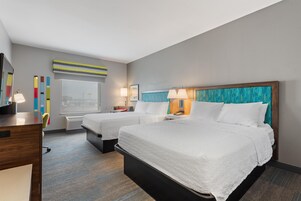 A hotel room with two beds, a desk, a chair, a window, and a wall with colorful artwork.