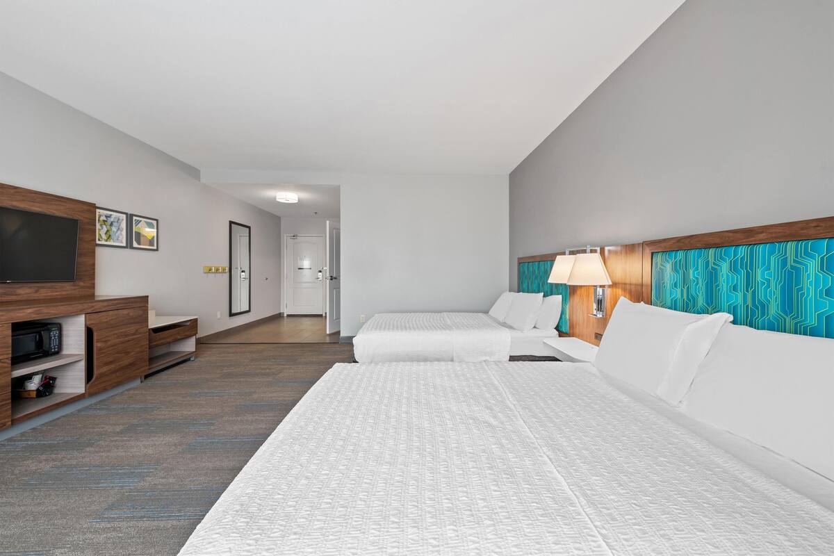A hotel room with a large bed, a TV, a desk with a lamp, and a view of the hallway.
