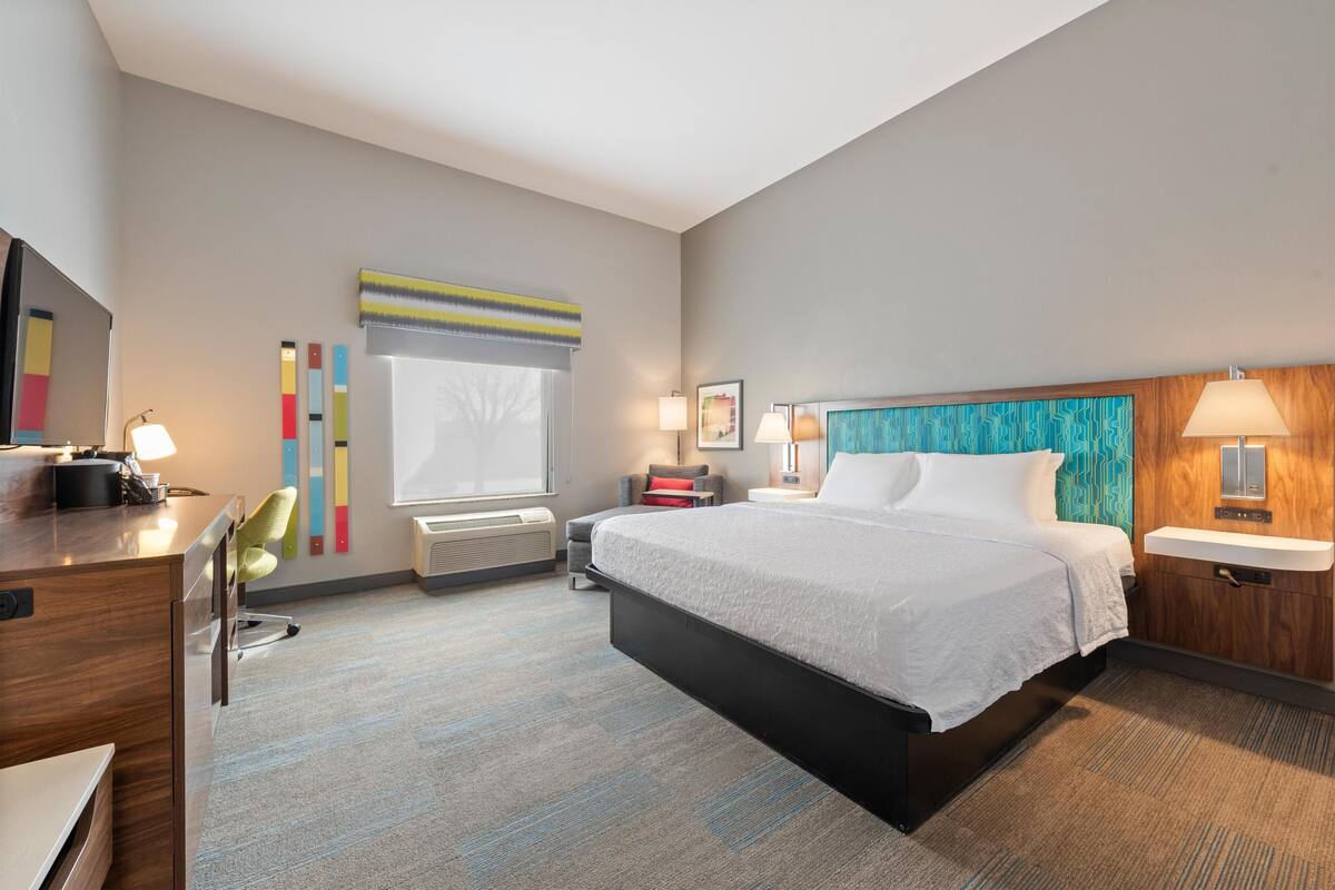 A hotel room with a bed, desk, chair, bedside table, lamp, and artwork on the wall.