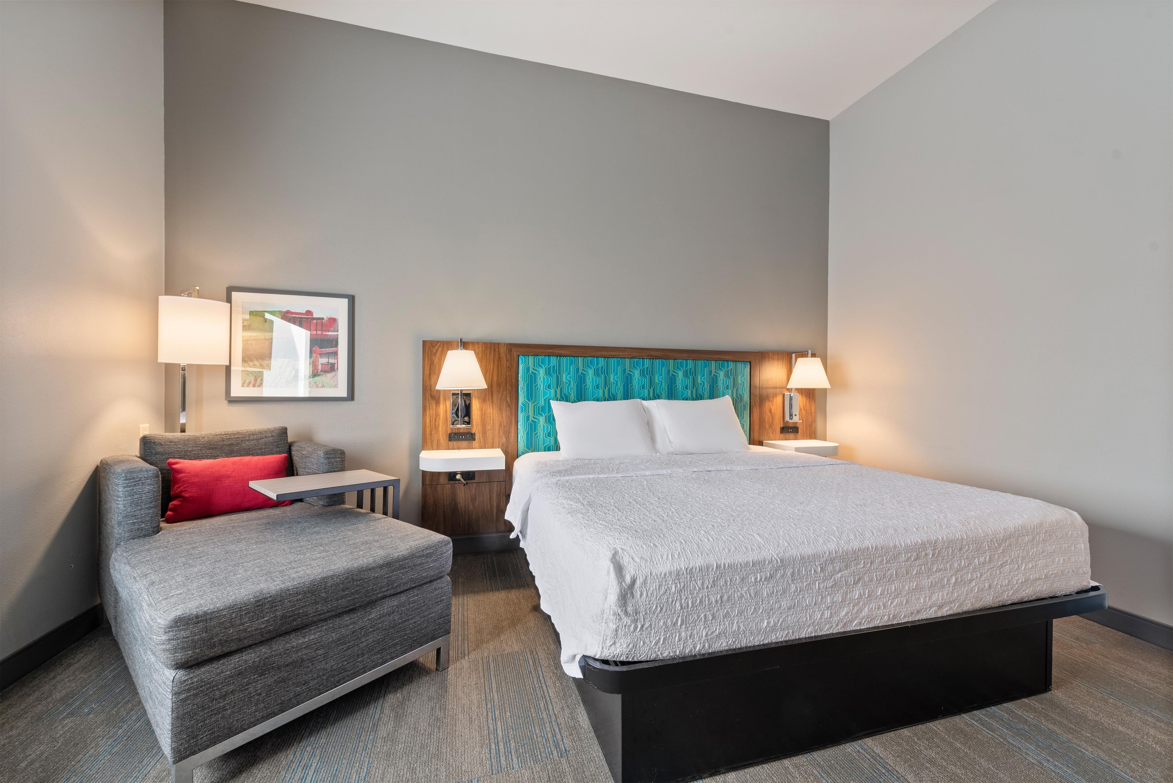 Una habitación de hotel con una cama, una silla, una mesita y un estante en la pared.