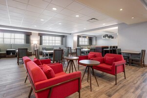 A modern hotel lobby with a mix of seating options, including red armchairs and grey sofas, a central dining area, and large windows providing natural light.