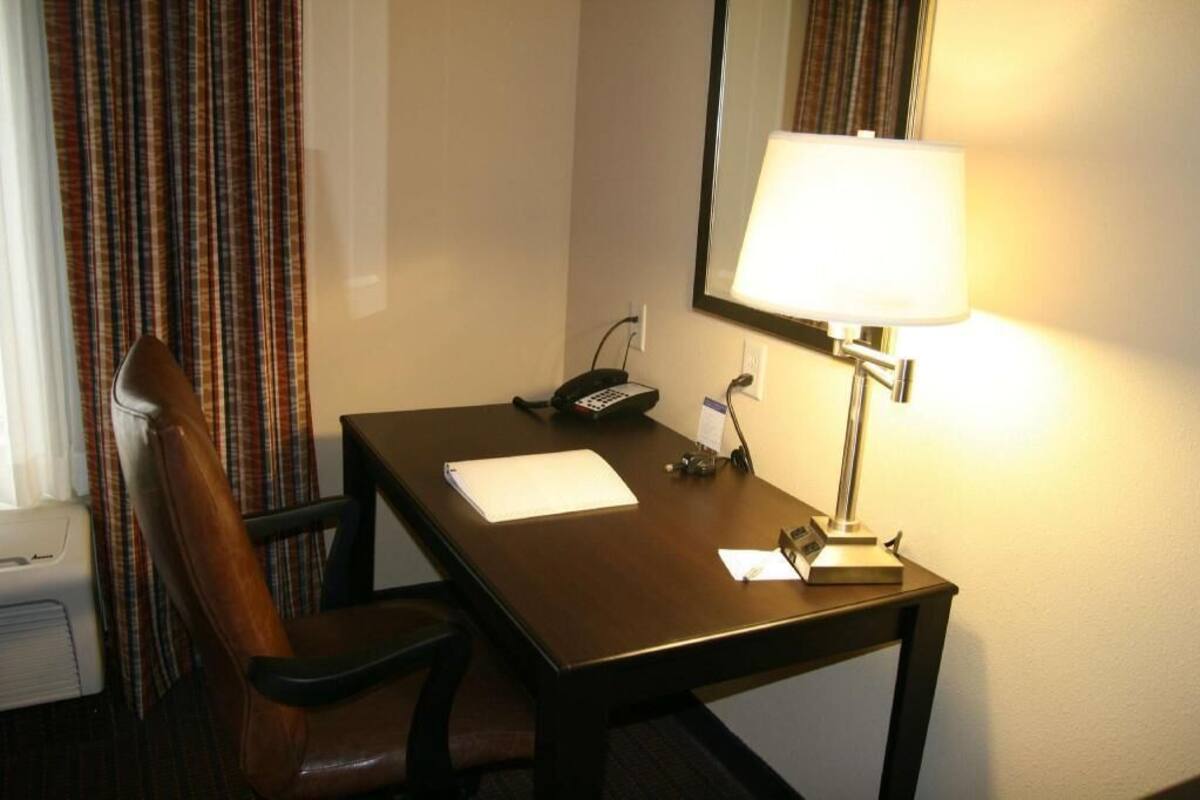 A hotel room with a wooden desk, a telephone, a lamp, a chair, and a window with curtains.