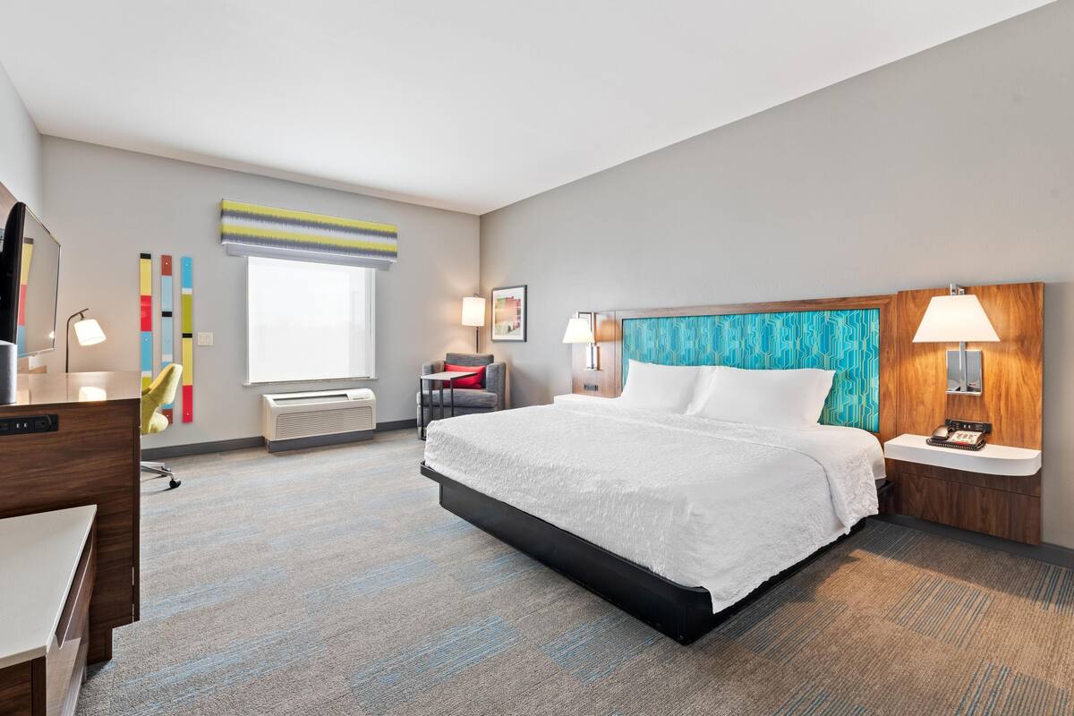 A hotel room with a large bed, bedside tables, a desk, a chair, a window with blinds, and a wall with a patterned design.