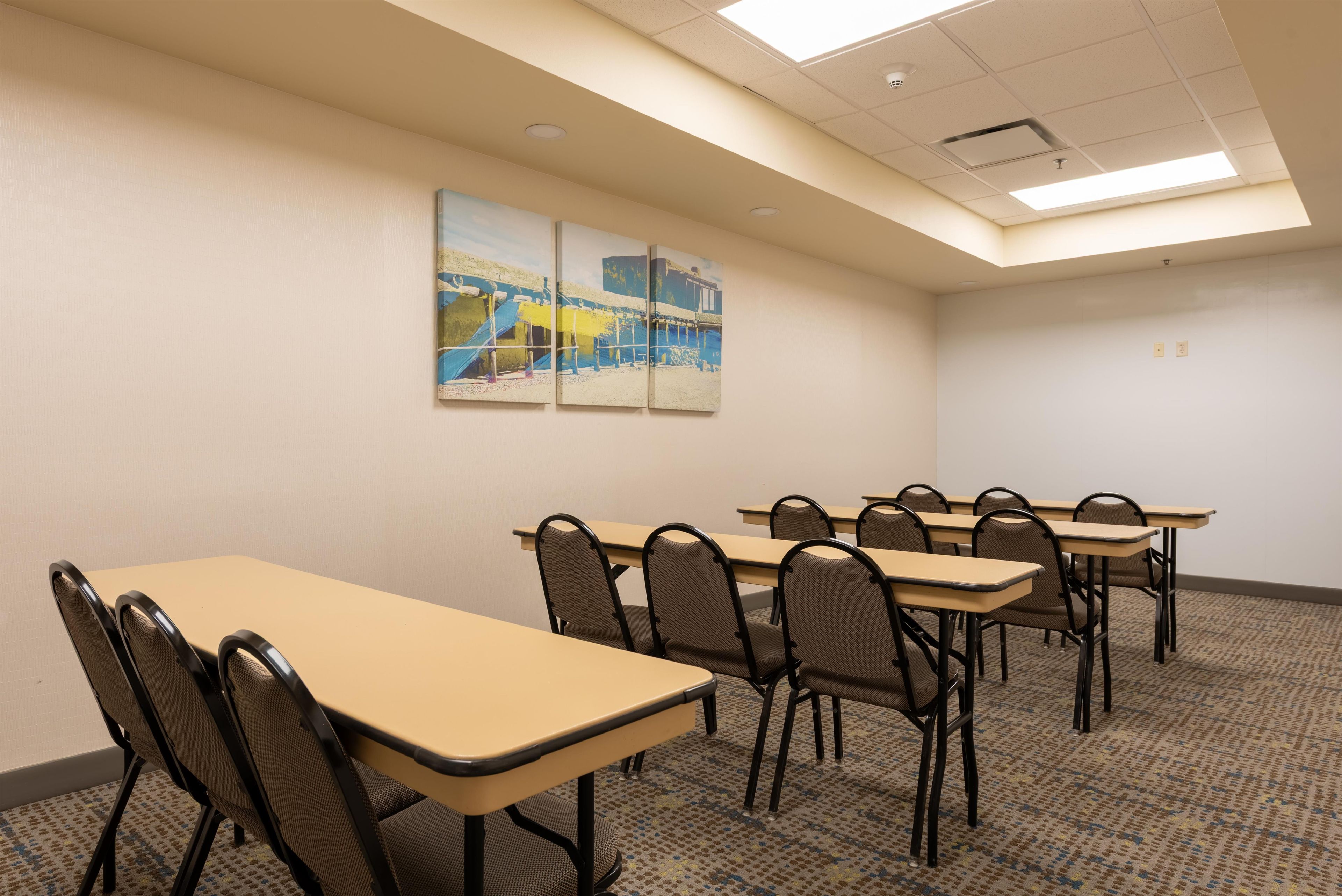 Une salle de conférence avec des tables et des chaises de couleur beige, un plafond doté d’un éclairage encastré et un tableau accroché au mur.