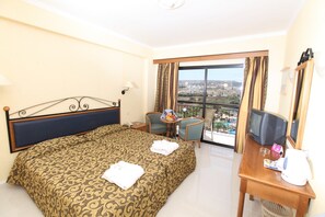A hotel room with a large bed, a desk with a television, and a balcony with a view of the city. - Tsokkos Gardens Hotel (Paralimni)