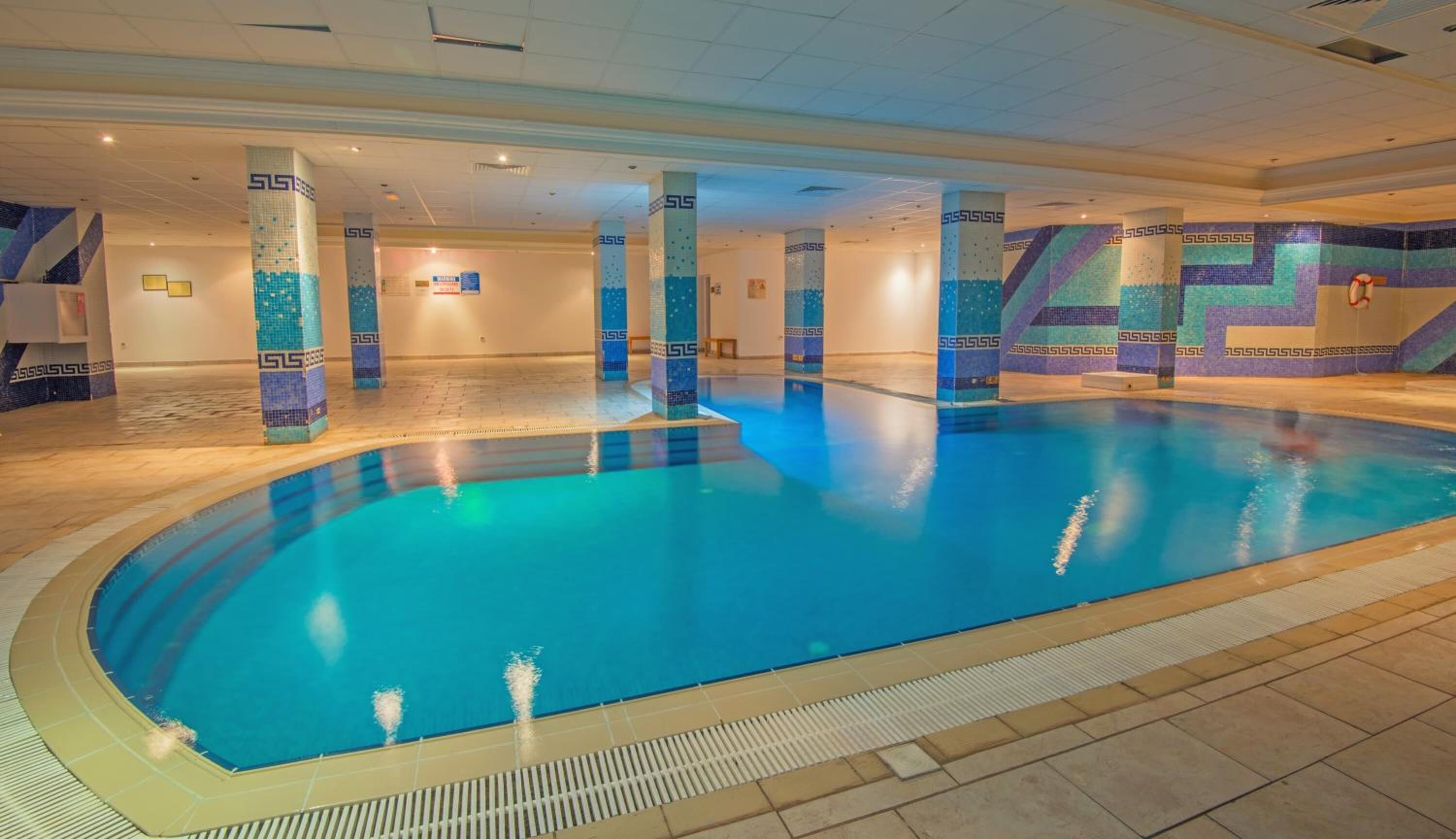 Une grande piscine intérieure, dont le sol et les murs sont recouverts de carreaux formant un motif géométrique, et qui est agrémentée de piliers portant le logo «  SLS ».
