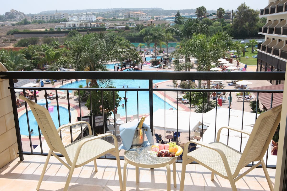 Un balcón con dos sillas de mimbre, una mesita con fruta y vistas a una piscina y zona de resort.