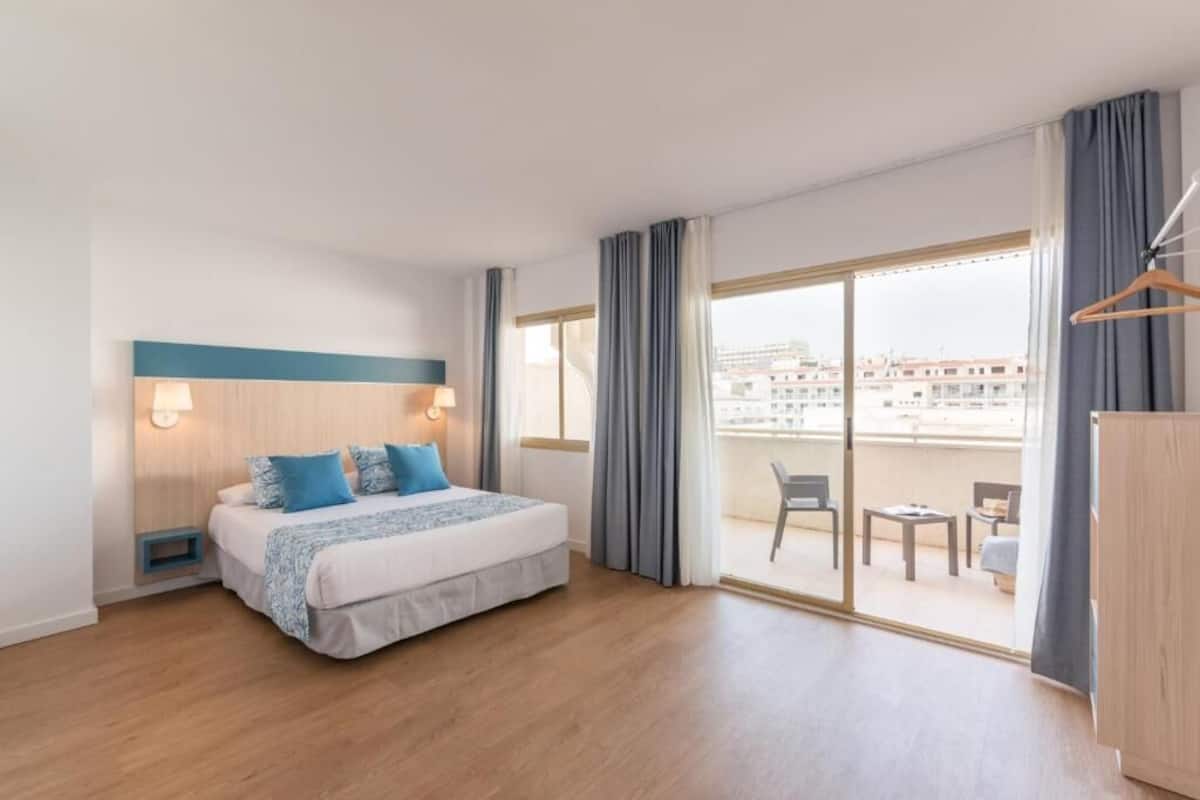 A hotel room with a bed, a desk, a chair, and a balcony with a view of buildings.