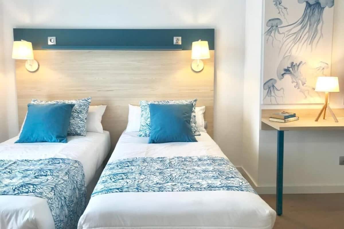 A hotel room with two beds, each with a blue and white patterned bedspread, a wooden headboard, and wall-mounted lamps.