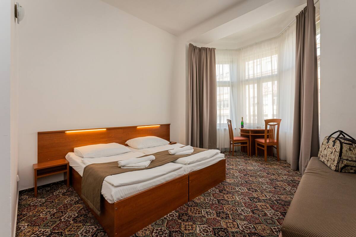 Habitación de hotel con una cama de madera, una mesita de noche pequeña, una alfombra estampada, un sofá, una mesa con sillas y una ventana con cortinas.