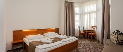 Habitación de hotel con una cama de madera, una mesita de noche pequeña, una alfombra estampada, un sofá, una mesa con sillas y una ventana con cortinas.
