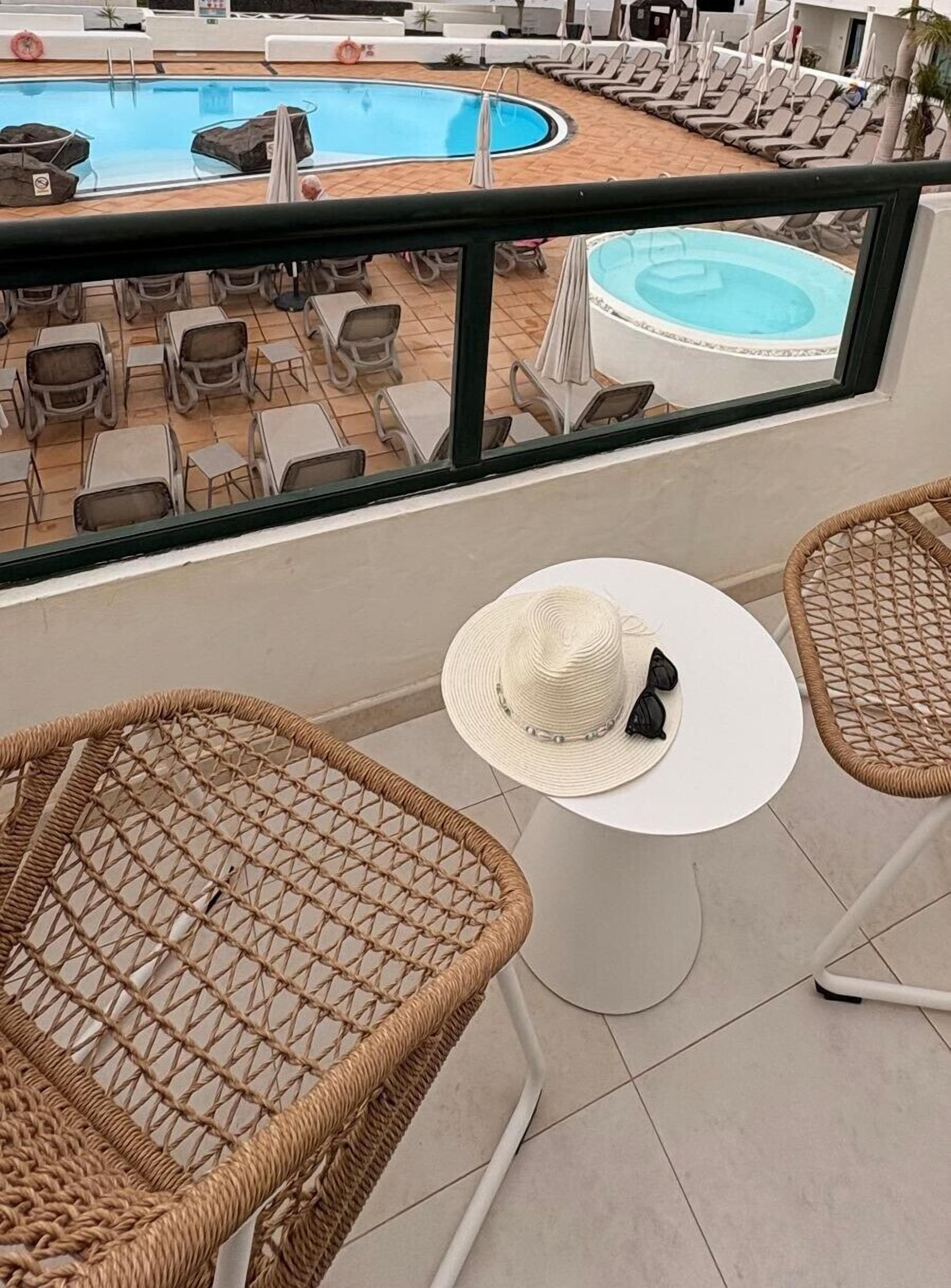 Un balcon avec des fauteuils en osier, une petite table sur laquelle reposent un chapeau et des lunettes de soleil, et un espace piscine avec des chaises longues et des parasols.