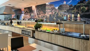 Área de buffet com mural de cachoeira, um chef preparando comida e variedade de frutas e bebidas.
