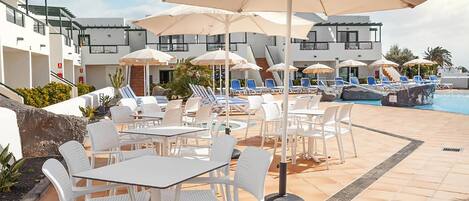 Een zwembadterras met witte ligbedden en tafels, parasols en een helder uitzicht op het zwembad.