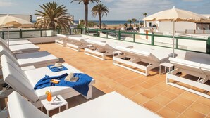Uma área de lounge no terraço com cadeiras e guarda-sóis brancos, piso ladrilhado e vista para a praia e palmeiras.