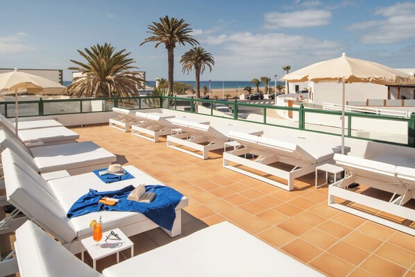 A rooftop lounge area with white lounge chairs and umbrellas, a tiled floor, and a view of the beach and palm trees.