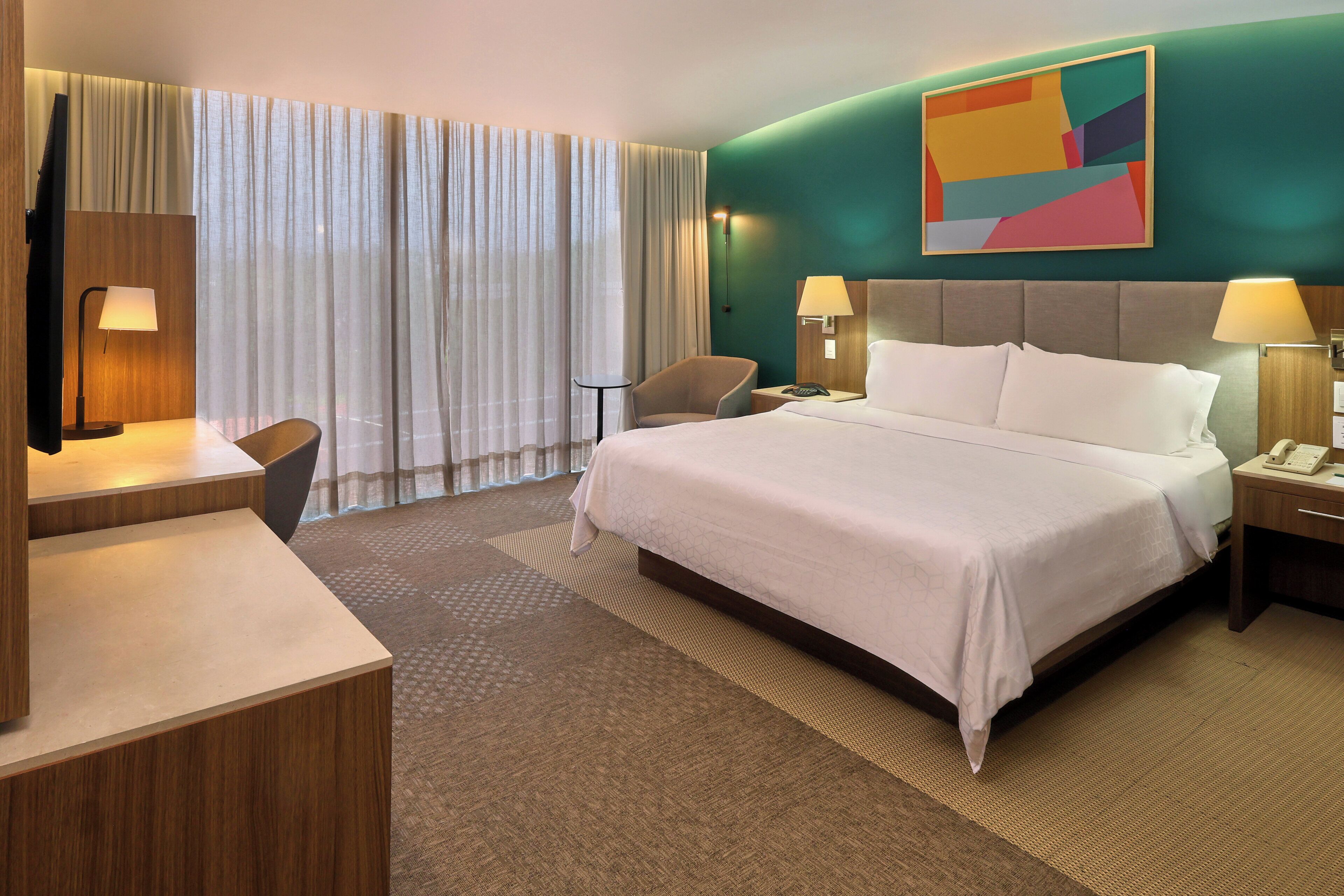 Ein Hotelzimmer mit Bett, Nachttischen, einem Schreibtisch, einem Sessel und einem großen Fenster mit Vorhängen.