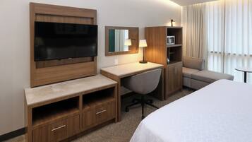 A hotel room with a bed, a desk, a chair, a TV, a lamp, and a minibar.