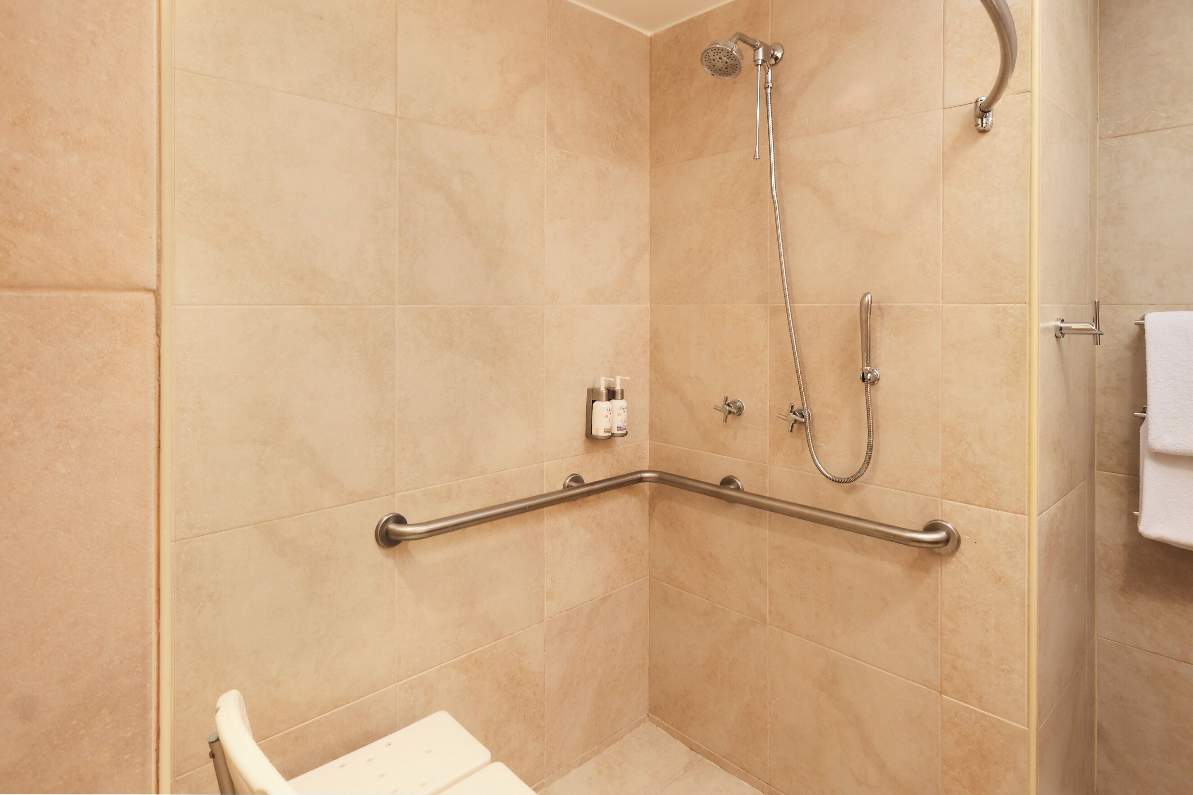 A bathroom with beige tiles, a shower with a handheld showerhead, a grab bar, and a white towel hanging on the wall.